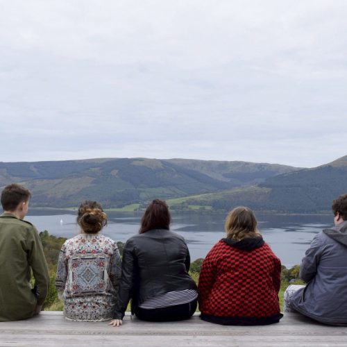 A group of young promoters who took part in the Get In programme. They are sitting outdoors in a line, with their backs to the camera. They are looking across a loch with hills beyond.