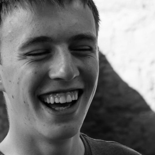 A close-up black and white image of a young man. He is smiling and laughing.