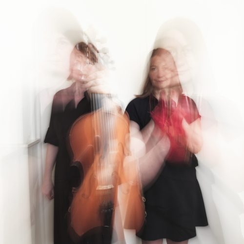 A blurred photo. Two women are seen, although not clearly. We can see that one woman is holding a fiddle, the other a pair of red shoes.