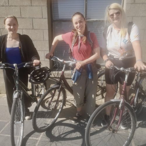 Penny Chivas and two others look towards camera, each holding a bicycle. Photo taken on the Burnt Out tour.
